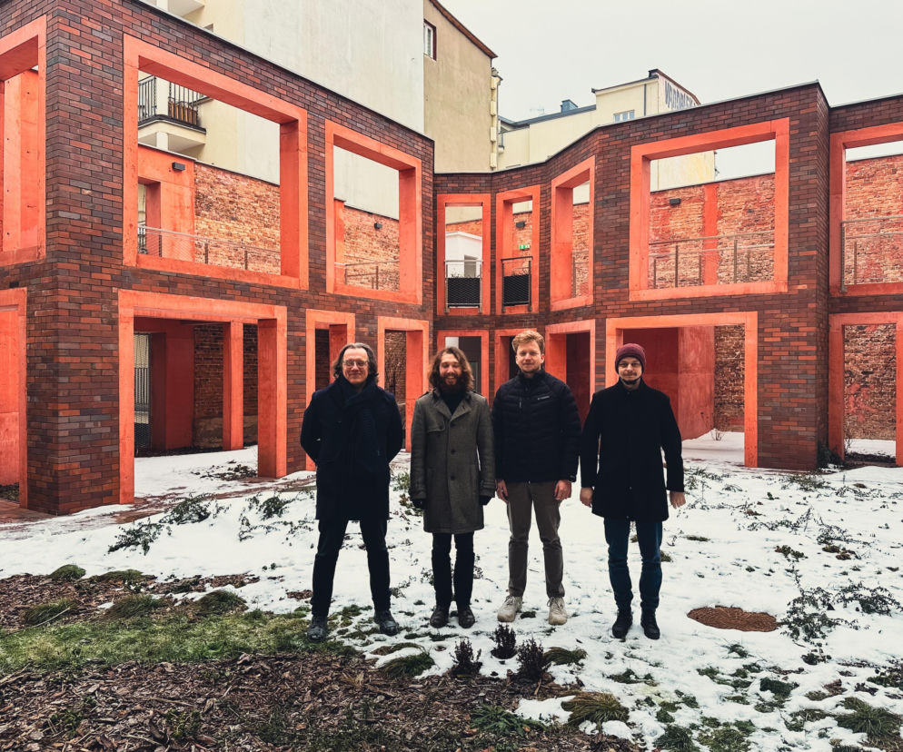 Czterech architektów z zespołu Grabczewscy Architekci, w tym Oskar Grąbczewski, stoi na ośnieżonym trawniku przed fasadą z cegły klinkierowej i intensywnie pomarańczowych ram. Architektura Murator Plus to portal, gdzie można dowiedzieć się więcej o ich projektach.