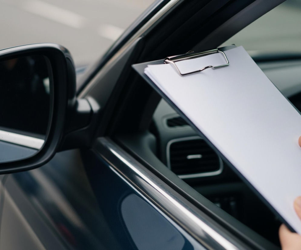 Dłonie trzymają czarną podkładkę z kilkoma białymi kartkami papieru przymocowanymi metalowym klipsem, umieszczone w otwartym oknie ciemnoniebieskiego samochodu. Samochód stoi na tle szarego, rozmytego otoczenia, a w jego wnętrzu widoczna jest kierownica i część deski rozdzielczej. Lewe lusterko boczne samochodu odbija jasne światło i fragmenty otoczenia.