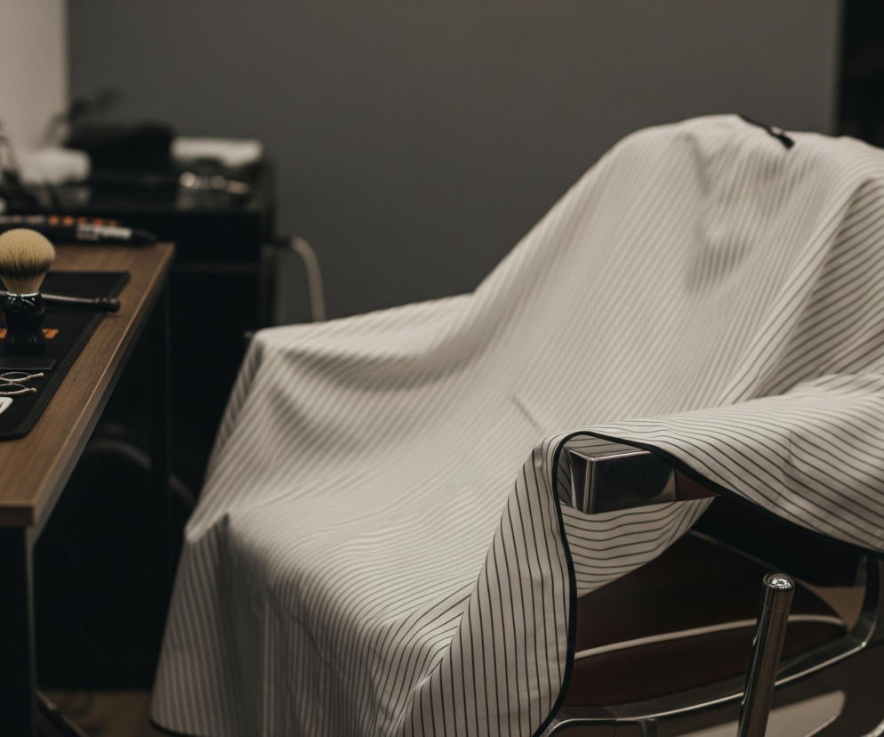 Krzesło fryzjerskie pokryte biało-czarną peleryną w paski zajmuje centralne miejsce, ustawione po prawej stronie. Po lewej stronie, na drewnianym blacie stołu, leży pędzel do golenia z jasnym włosiem i ciemną rączką, obok niego widać nożyczki i grzebienie ułożone na czarnej podkładce. W tle, w rozmyciu, stoją białe i czarne butelki z pompkami, a ściany są w odcieniach szarości.