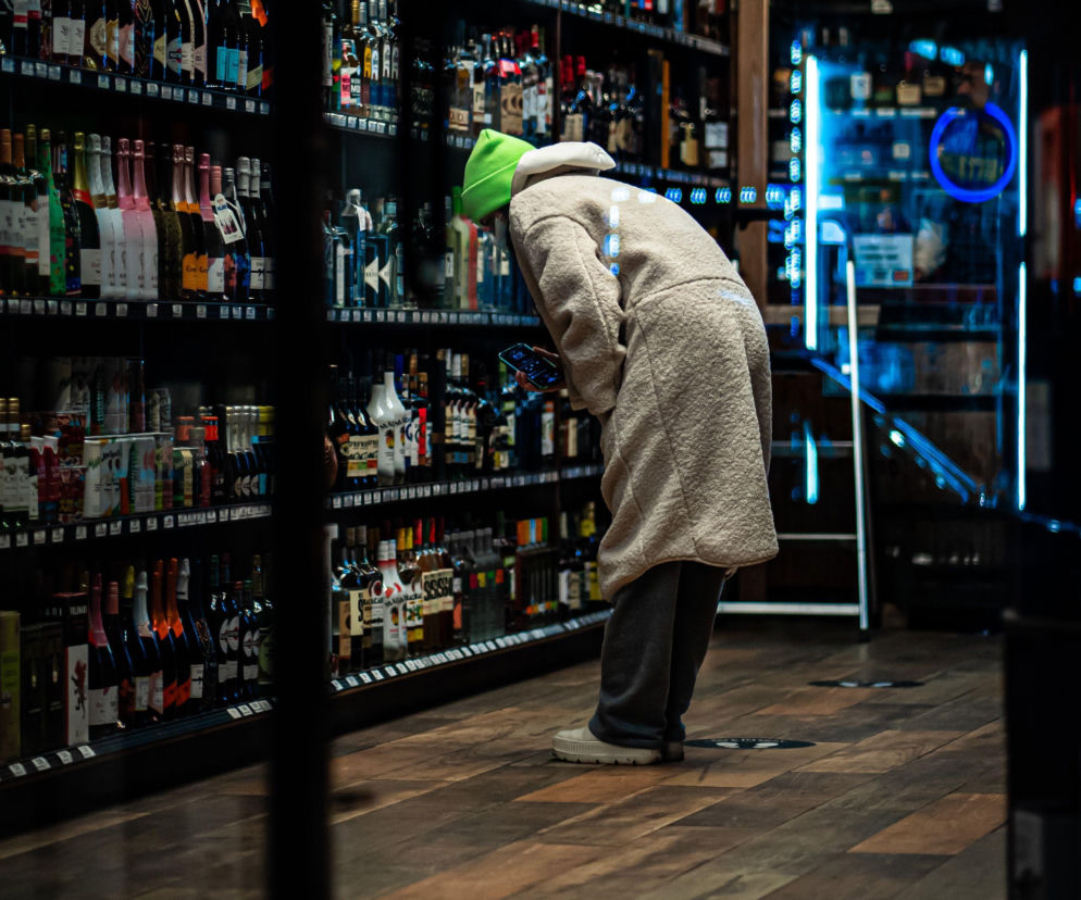 Osoba w zielonej czapce wybiera alkohol na regale sklepowym. O nowej opłacie od używek przeczytasz na Eska.