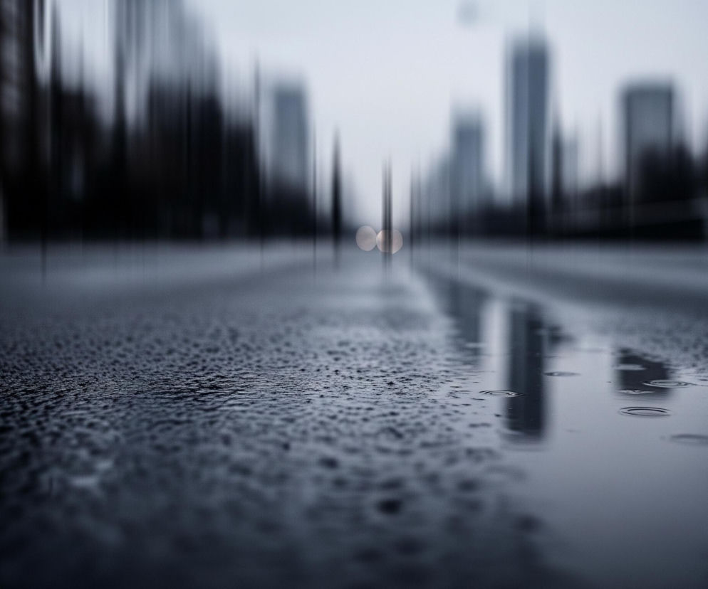 Ciemna, mokra droga lub chodnik pokryta jest licznymi kroplami wody w dolnej części obrazu, tworzącą kałużę po prawej stronie, w której odbija się rozmyty, pionowy obiekt. Tło przedstawia rozmyte kontury wysokich budynków miejskich w odcieniach szarości i błękitu, z kilkoma jasnymi, okrągłymi punktami światła widocznymi w oddali na środku. Cała scena utrzymana jest w chłodnych, ciemnych tonacjach.