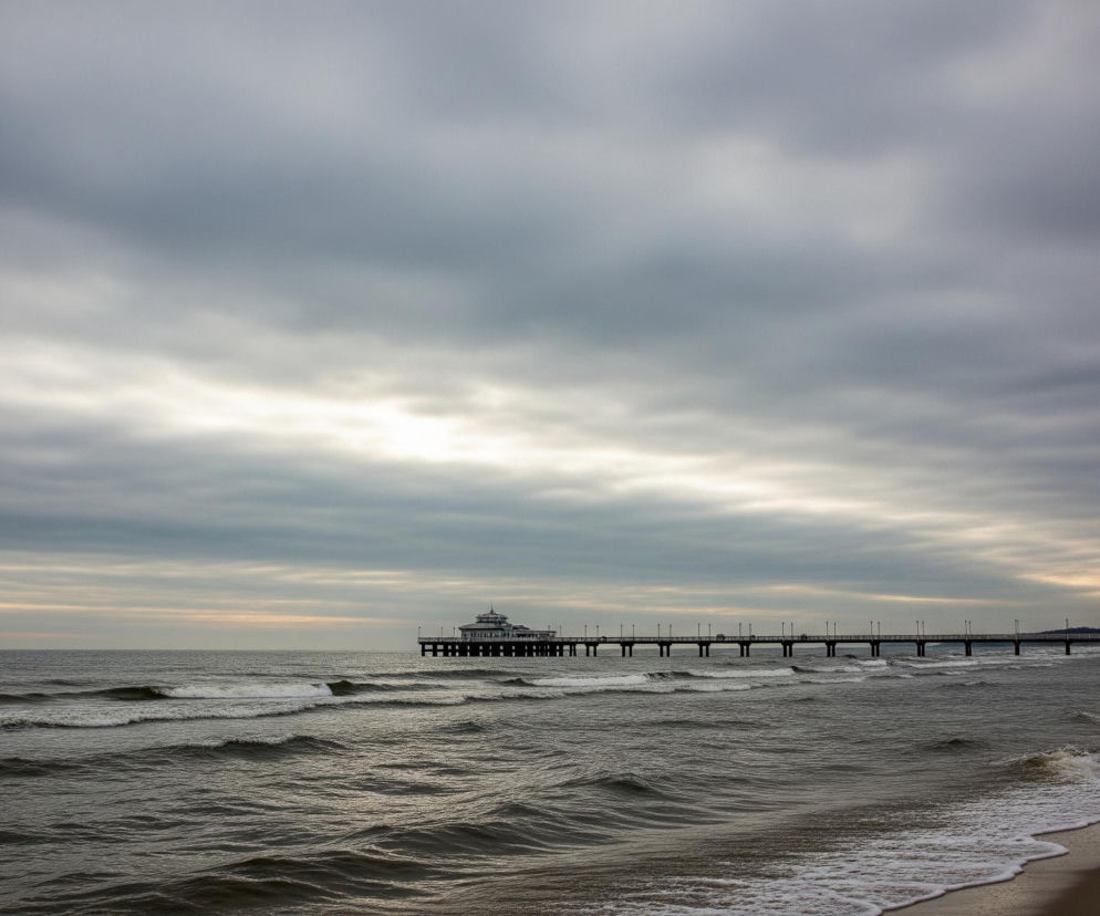 Na zdjęciu dominuje pochmurne, szare niebo z jaśniejszym pasmem wzdłuż horyzontu. Poniżej rozciąga się morze o ciemnych, falujących wodach, z widocznymi białymi grzywami fal przy brzegu, gdzie piasek jest ciemnożółty. Długie molo z licznymi latarniami rozciąga się w głąb morza, prowadząc do biało-zielonego budynku z charakterystycznym, okrągłym dachem. W oddali, na plaży widać sylwetki kilku osób, a także zarys zabudowań i drzew na linii brzegowej.