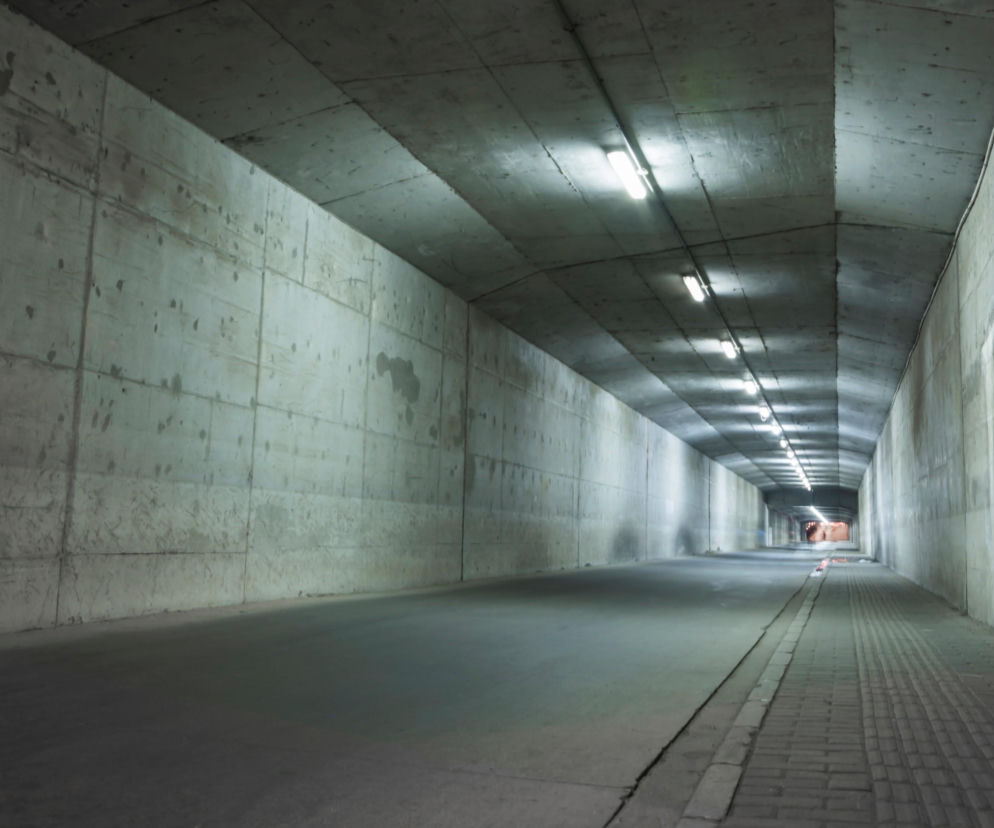 Pusty, betonowy tunel z rzędem świetlówek na suficie, oświetlających drogę i chodnik. Obiekt symbolizuje infrastrukturę ochronną dual-use, o której więcej przeczytasz na naszym portalu, podkreślając potrzebę budowy systemów odporności.