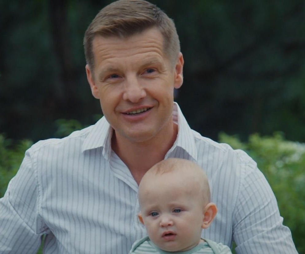 Rafał Mroczek jako Paweł Zduński z uśmiechem trzyma na rękach niemowlę, Antosia, ubranego w zielone body, na tle zielonych krzewów, co zapowiada zmiany w serialu M jak miłość. Więcej o tym przeczytasz na stronie Super Seriale.