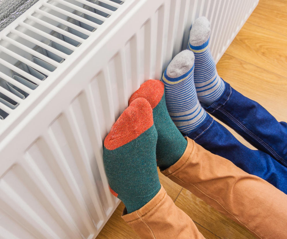 Dwie osoby w grubych skarpetkach grzeją stopy przy kaloryferze. O sposobach na ciepło w domu przeczytasz w Eska.