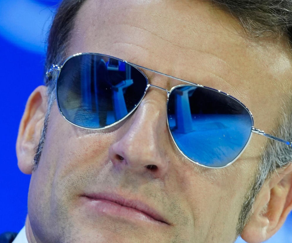 Emmanuel Macron w okularach przeciwsłonecznych typu aviator podczas wystąpienia, z widocznym zarostem i odblaskami błękitu na szkłach. Temat okularów Macrona i ich wpływu na rynek finansowy znajdziesz na Super Biznes.