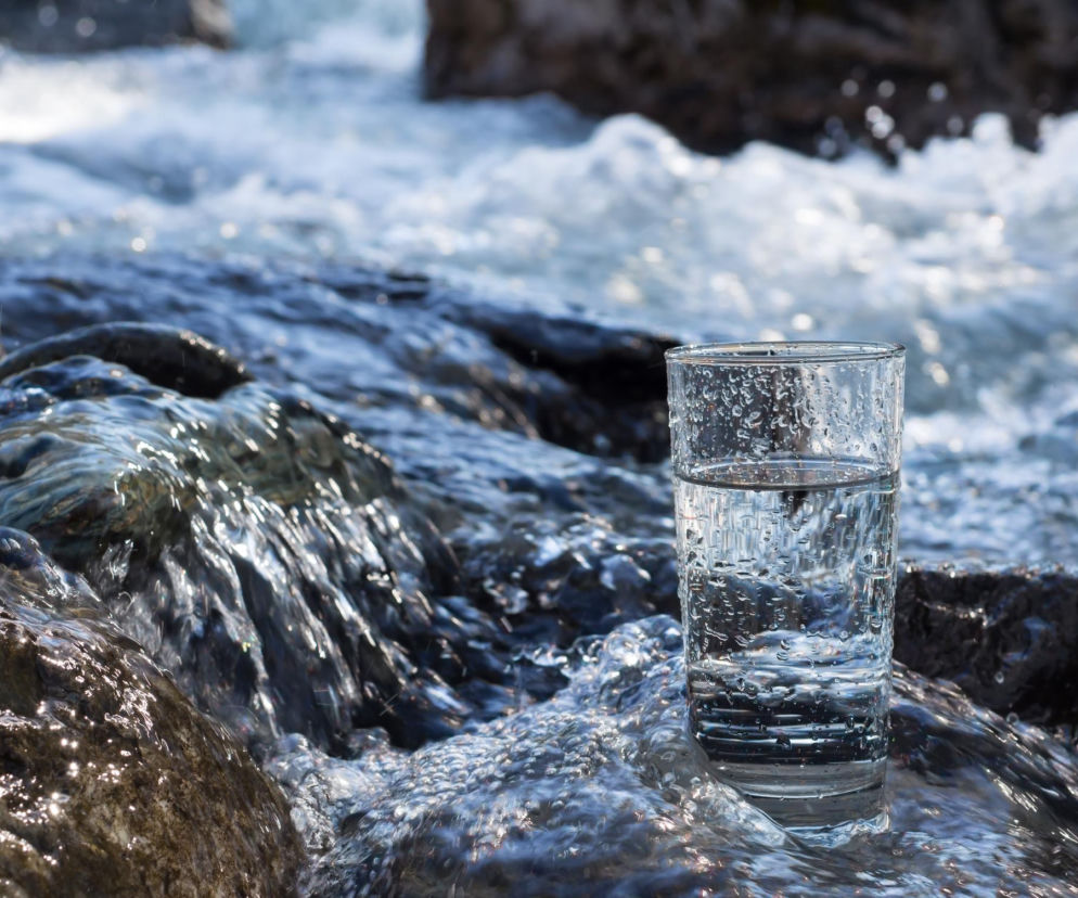 Szklanka wypełniona czystą, świeżą wodą, stojąca na kamieniu pośród strumienia, z bąbelkami i kroplami wody na szkle, symbolizująca naturalne źródło i świeżość. Artykuł na Poradnik Zdrowie wyjaśnia kryteria klasyfikacji wód mineralnych.