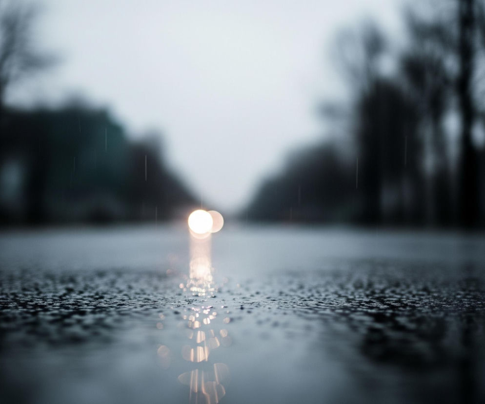 Droga pokryta jest drobnymi kropelkami wody, co sugeruje deszcz; powierzchnia jest ciemna, a jej faktura przypomina asfalt. W oddali, na końcu drogi, widoczne są dwa jasne, rozmyte punkty światła, odbijające się długą, świetlistą smugą w wodzie na nawierzchni. Po obu stronach drogi rozciągają się niewyraźne, ciemne sylwetki drzew lub budynków, rozmywające się w szarym, rozmytym tle nieba. Ogólna kolorystyka obrazu jest zimna, z dominującymi odcieniami szarości i granatu, z wyraźnym akcentem ciepłego światła w tle.