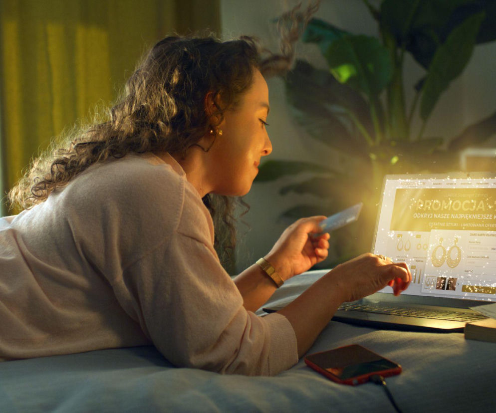 Kobieta na łóżku kupuje biżuterię przez laptopa z napisem promocja. O cyberbezpieczeństwie przeczytasz na SE.