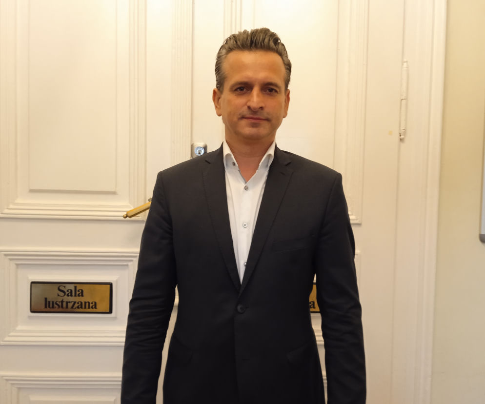 Poseł Robert Wardzała w garniturze stoi przed drzwiami do Sali Lustrzanej. O polityku przeczytasz na SE.