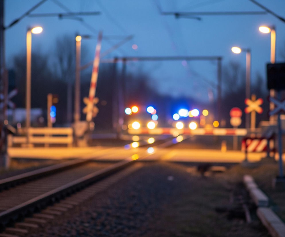 Na pierwszym planie widoczne są dwa tory kolejowe, ciągnące się przez środek kadru, ułożone na szarym żwirze. W tle, na środku obrazu, znajduje się rozmyty przejazd kolejowy z opuszczonymi szlabanami w biało-czerwone pasy oraz kilkoma świecącymi, pomarańczowymi latarniami ulicznymi po obu stronach. Dalej, za przejazdem, widoczne są rozmyte światła pojazdu – pomarańczowe i niebieskie, sugerujące obecność pociągu lub innego pojazdu uprzywilejowanego. Całość obrazu utrzymana jest w ciemnych, nocnych barwach, z dominującym niebieskim i pomarańczowym odcieniem świateł.
