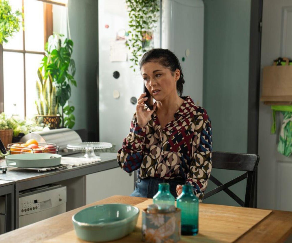 Katarzyna Cichopek jako Kinga Zduńska w kuchni, rozmawiająca przez telefon, z zatroskaną miną. W tle okno z roślinami i lodówka. Scena z serialu M jak miłość w TVP2, o której przeczytasz więcej na portalu Super Seriale.