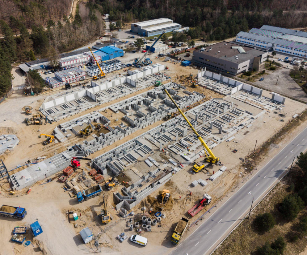 Obszerny plac budowy, widok z lotu ptaka, z konstrukcjami fundamentów magazynów energii PGE, ciężkim sprzętem budowlanym oraz drogą asfaltową obok. Dowiedz się więcej o strategicznych inwestycjach PGE w magazyny energii, które wzmocnią bezpieczeństwo energetyczne kraju na naszym portalu.