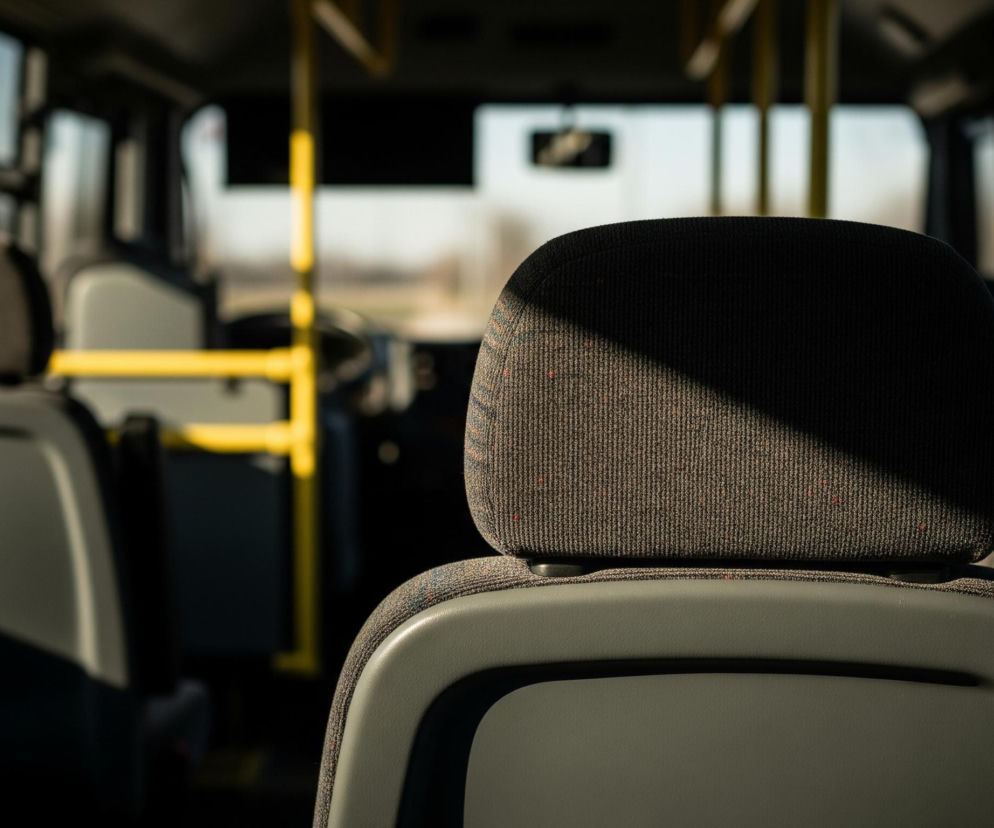Wnętrze autobusu wypełnione jest ciemnymi siedzeniami, z których jedno znajduje się na pierwszym planie, częściowo zacienione. Siedzenia po lewej stronie są podobne, ale jaśniejsze, bardziej widoczne. W tle rozciąga się przestrzeń autobusu z żółtymi poręczami i jasnym, rozmazanym widokiem zza przedniej szyby. Światło wpada przez okna, tworząc wyraźne kontrasty światłocieni.