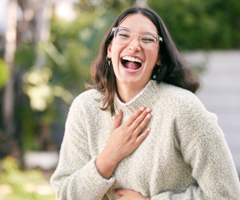 Roześmiana kobieta w okularach, trzymająca ręce na klatce piersiowej i brzuchu, śmieje się głośno. To obraz, który idealnie ilustruje temat niemożności samodzielnego połaskotania się, o którym pisze Poradnik Zdrowie. Mózg ignoruje własny dotyk, aby oszczędzić zbędnych reakcji.