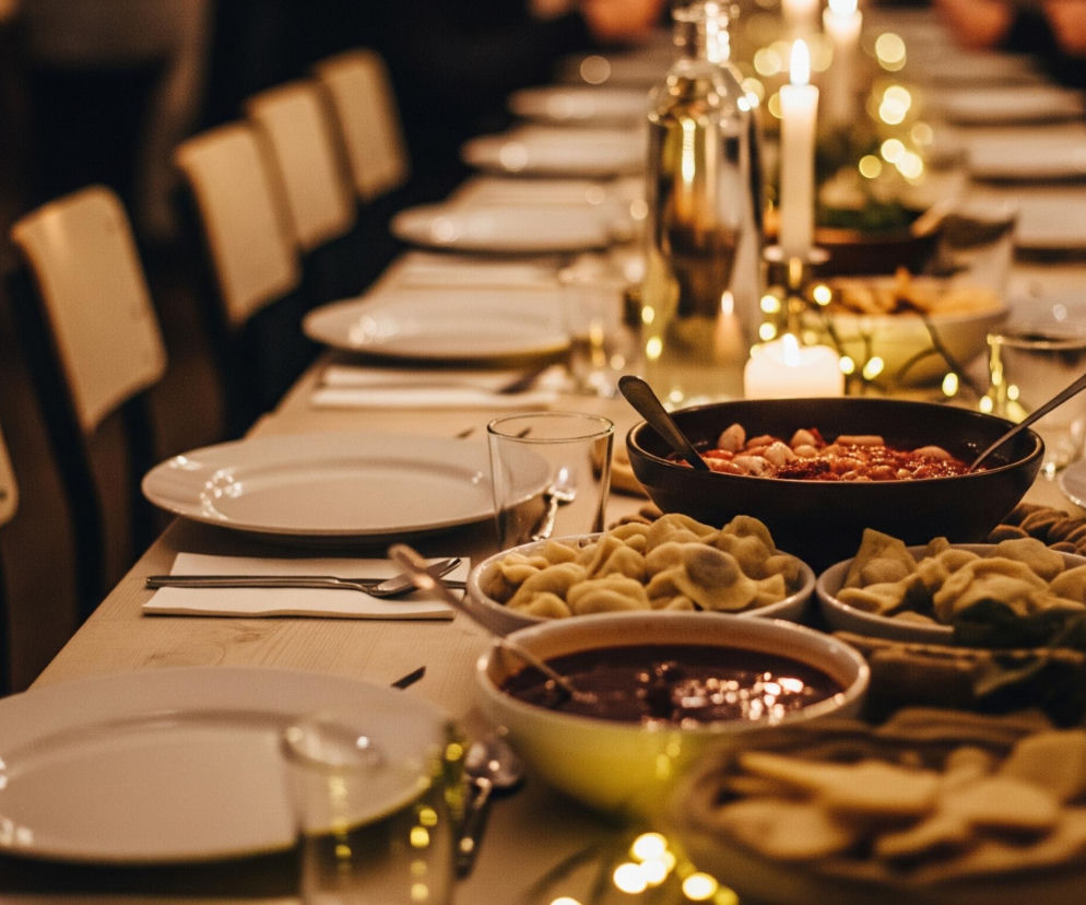 Długi stół biesiadny jest centralnym punktem obrazu, zastawiony licznymi daniami i zastawą. Na blacie widać białe talerze, szklanki i sztućce ułożone na serwetkach, przygotowane dla wielu osób. Wzdłuż stołu stoją miski z jedzeniem, w tym duża brązowa misa z czerwoną potrawą z kluskami oraz kilka mniejszych białych miseczek z pierogami i czerwoną pastą lub sosem. Pomiędzy potrawami rozmieszczone są zapalone świece w świecznikach oraz girlandy ze złotymi, rozświetlonymi lampkami, które nadają ciepły, świąteczny nastrój. Tło stanowi rząd krzeseł z jasnymi oparciami oraz rozmazane sylwetki ludzi siedzących przy stole, co sugeruje spotkanie towarzyskie lub uroczysty posiłek. Dominują ciepłe barwy, głównie brązy, beże i złote odcienie świateł, a oświetlenie jest przyćmione.