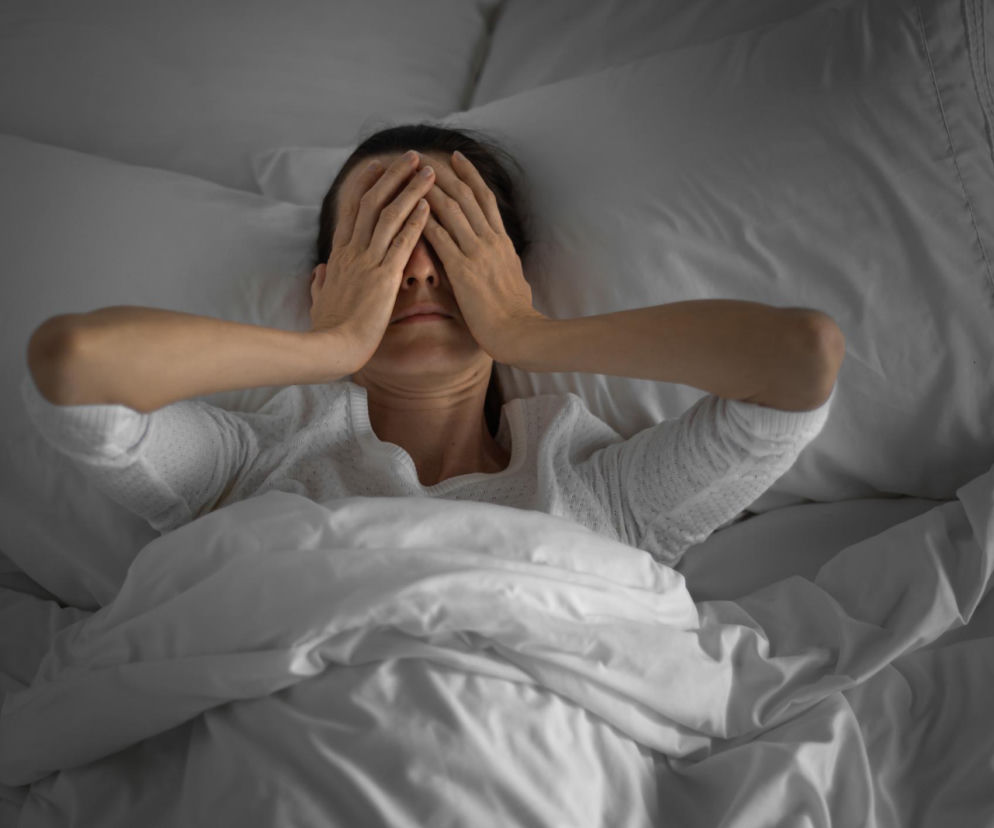 Kobieta leżąca w łóżku z twarzą zasłoniętą dłońmi, symbolizująca nocne poty i niepokój związany z mokrą pościelą. Zdjęcie ilustruje problem nadmiernego pocenia się w nocy i konieczność poznania jego przyczyn, o czym przeczytasz na Poradnik Zdrowie.