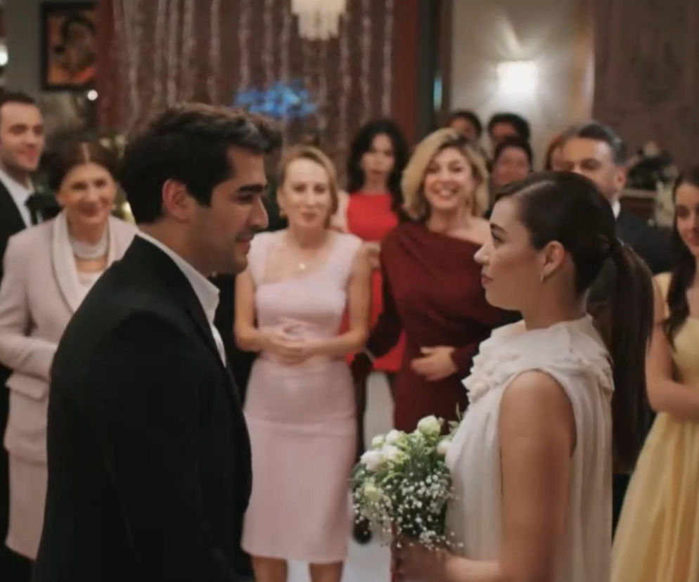 Mert Ramazan Demir (Ferit) i Afra Saracoglu (Seyran) podczas ceremonii ślubnej w tureckim serialu Złoty chłopak. Para stoi naprzeciwko siebie, Seyran w białej sukni z bukietem, Ferit w garniturze, a w tle widać uśmiechniętych gości, co sugeruje radosną scenę z serialu dostępnego na Super Seriale.