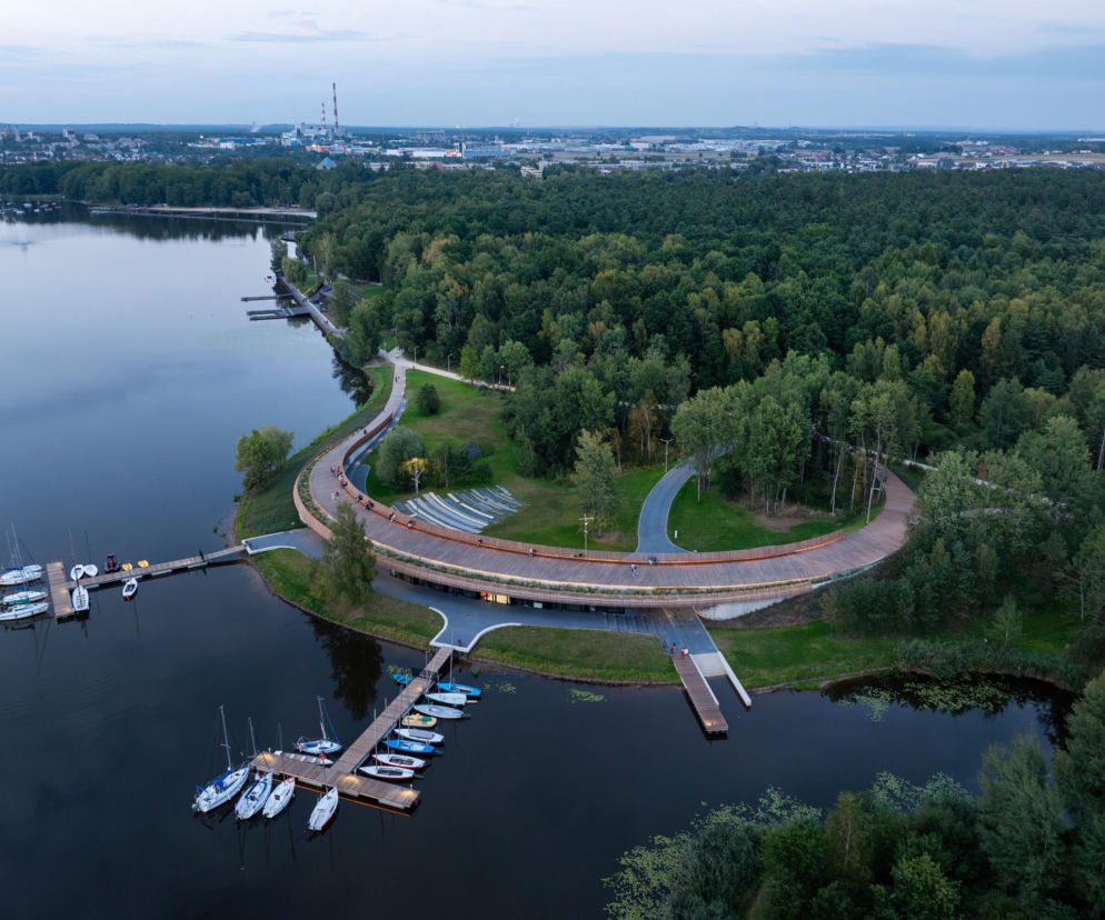 Widok z lotu ptaka na Przystań Żeglarską na Jeziorze Paprocańskim w Tychach. Zdjęcie ukazuje drewnianą promenadę, nowe pomosty z zacumowanymi łodziami oraz otaczającą zieleń, w tle panoramę miasta. Więcej o tym na Architektura Murator Plus.