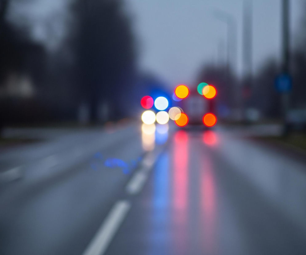 Rozmyty obraz przedstawia ulicę z poruszającymi się pojazdami, widzianą z perspektywy kierowcy. Na pierwszym planie widać ciemnoszarą, mokrą nawierzchnię drogi z rozmytą białą linią przerywaną po lewej stronie, a także słabo widoczne, rozmyte odbicia świateł na jej powierzchni. W oddali, w centrum kadru, znajdują się jaskrawe, kolorowe plamy światła, reprezentujące reflektory i tylne światła pojazdów, w tym niebieskie, czerwone, białe i pomarańczowe. Tło stanowi niewyraźny krajobraz z ciemnymi kształtami drzew i słupów, otoczony szarym, zamglonym niebem.