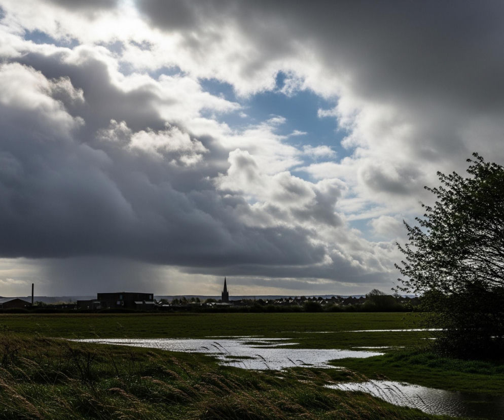 W górnej części kadru dominują dramatyczne, ciemne chmury burzowe, z prześwitami jasnego, błękitnego nieba i słońca w centrum, tworząc efektowny kontrast. Poniżej rozciąga się zielona, podmokła łąka z licznymi kałużami odbijającymi światło, szczególnie widocznymi w lewej i środkowej części zdjęcia. Na horyzoncie widoczna jest sylwetka małego miasteczka lub wsi z charakterystycznym, spiczastym dachem kościoła lub wieży po prawej stronie, a także budynkami i drzewami w oddali. Po prawej stronie kadru, na pierwszym planie, rośnie krzew o ciemnych, gęstych liściach, z gałęziami dynamicznie ułożonymi w stronę lewej.