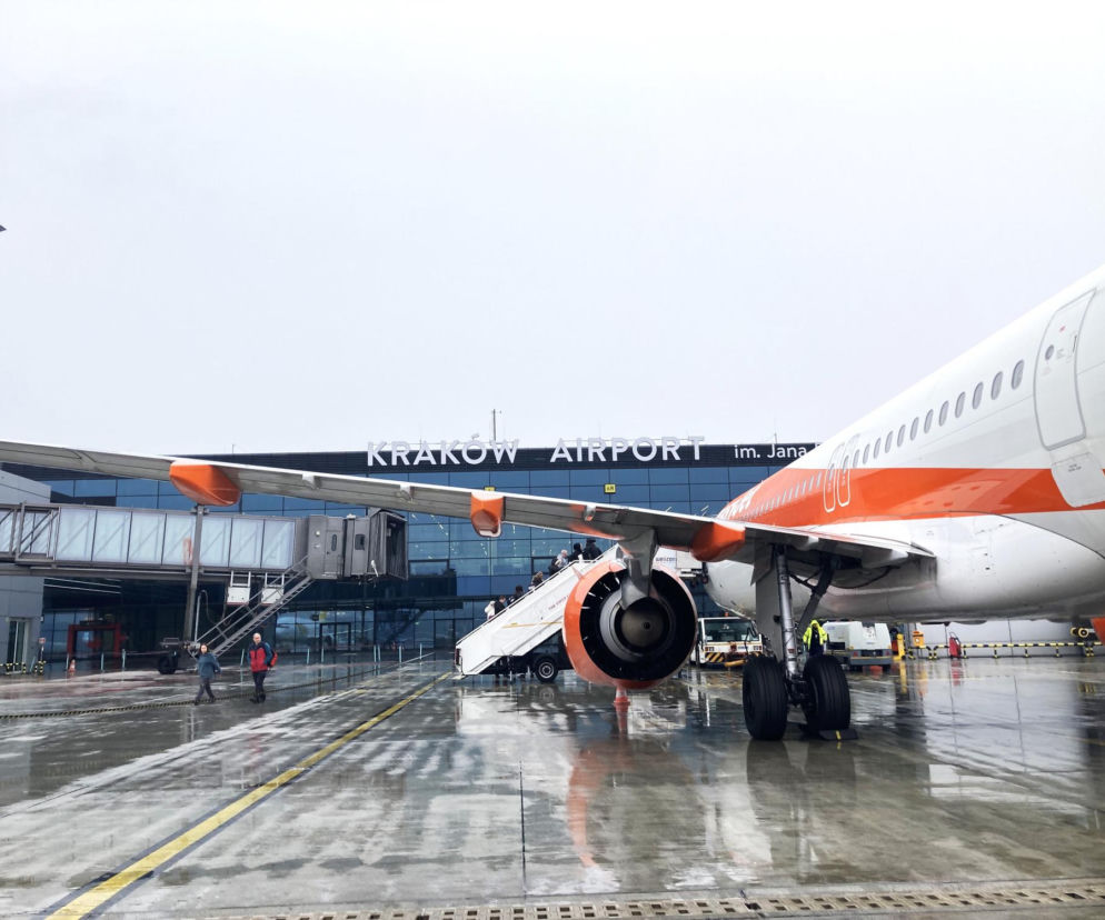 Biało-pomarańczowy samolot na mokrej płycie lotniska Kraków Airport. O inwestycjach w Balicach przeczytasz na Architektura.