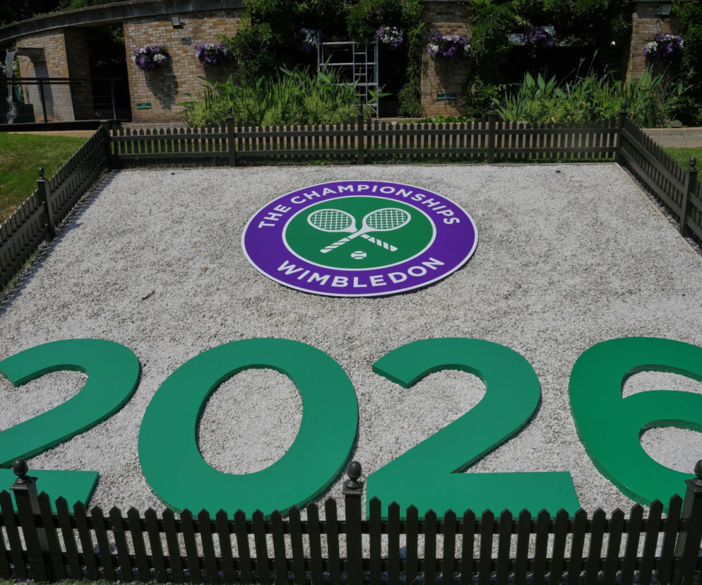 Logotyp Wimbledon Championships z rakietami i piłką tenisową, a pod nim zielone cyfry 2026, oznaczające rok turnieju, otoczone drewnianym płotem. W tle zieleń i fragmenty zabudowań. Więcej o Wimbledonie na naszym portalu.