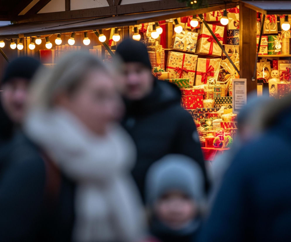 Centralnym elementem jest oświetlony stragan, wypełniony licznymi, kolorowymi, zapakowanymi prezentami, głównie w odcieniach czerwieni, złota, zieleni i bieli, przewiązanymi wstążkami. Nad ladą wisi rząd jasnych, białych żarówek, rzucających ciepłe światło na towary, a na dachu widoczne są drobne lampki choinkowe. Na pierwszym planie, rozmyte postacie ludzi, w tym kobieta w jasnym szaliku oraz osoby w ciemnych czapkach i kurtkach, przechodzą obok straganu. Tło jest ciemne, sugerując późną porę lub zmierzch.