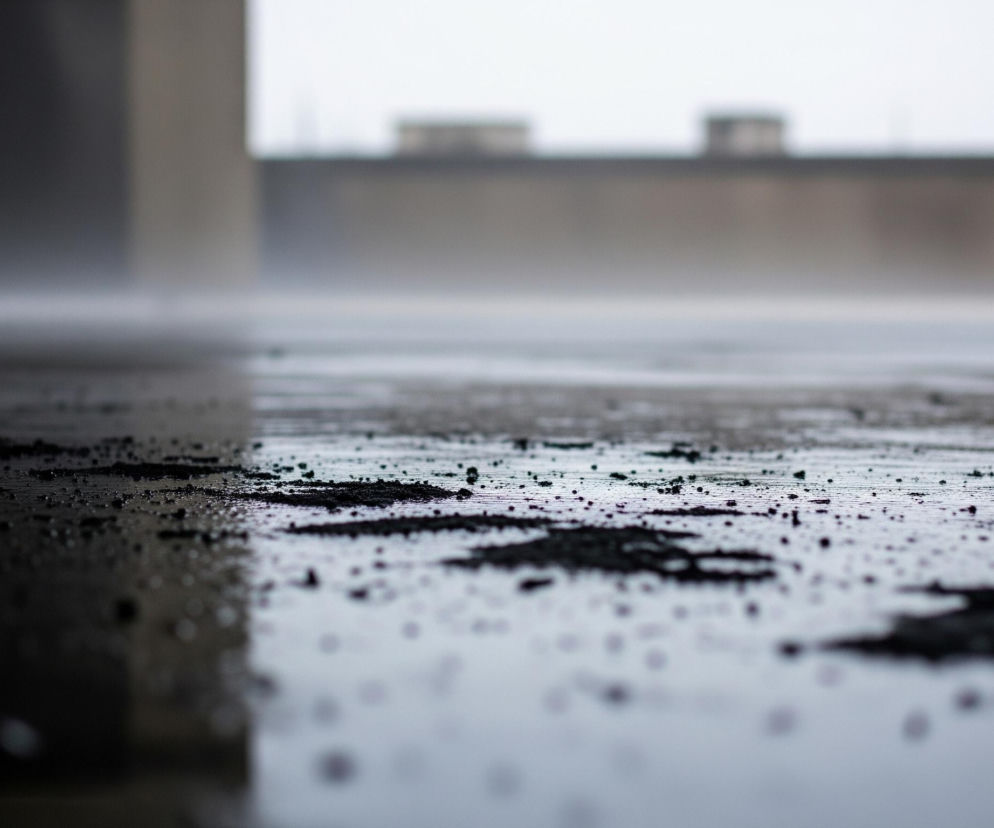Mokra, szara powierzchnia, prawdopodobnie podłoga, rozciąga się od dołu obrazu, pokryta licznymi drobnymi kroplami wody i większymi, nieregularnymi plamami ciemnego, rozmazanego materiału. Lewa strona pierwszego planu jest ciemniejsza, tworząc kontrast z jaśniejszym obszarem po prawej. W tle widać zamglony, szary horyzont z niewyraźnymi, prostokątnymi kształtami budynków lub konstrukcji. Całość utrzymana jest w stonowanej kolorystyce szarości, bieli i czerni, tworząc atmosferę wilgoci i spokoju.