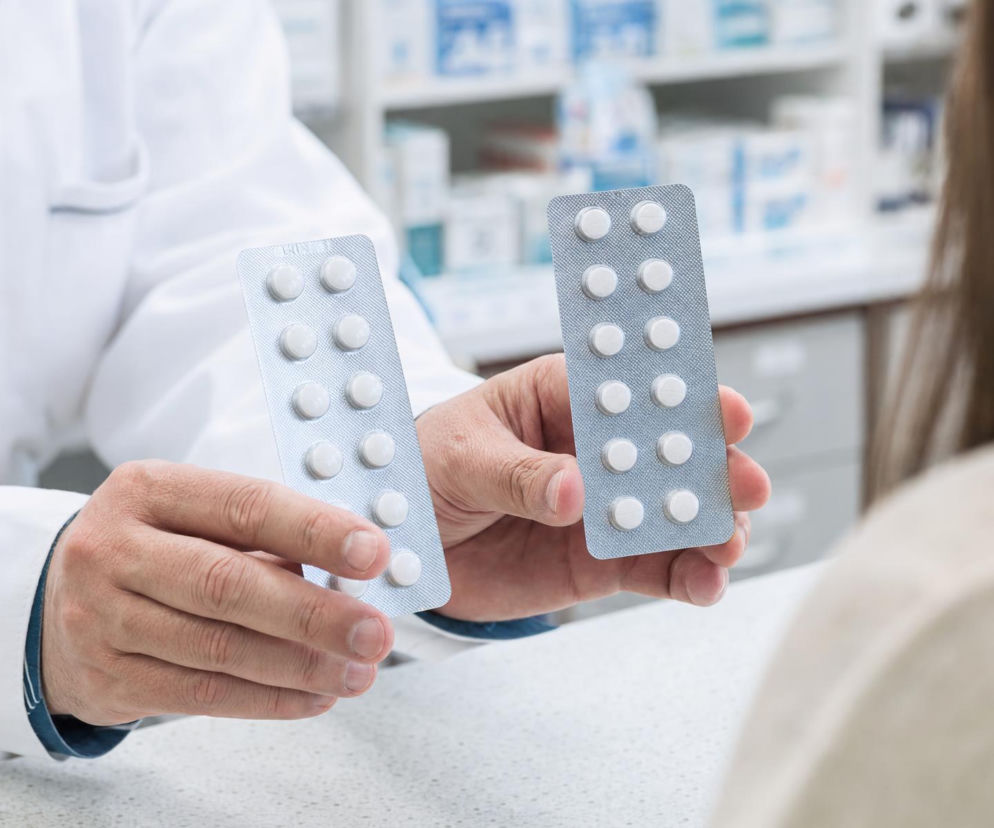 Dłonie farmaceuty w białym fartuchu trzymają dwa blistry z białymi tabletkami. O lekach generycznych przeczytasz na PZ.