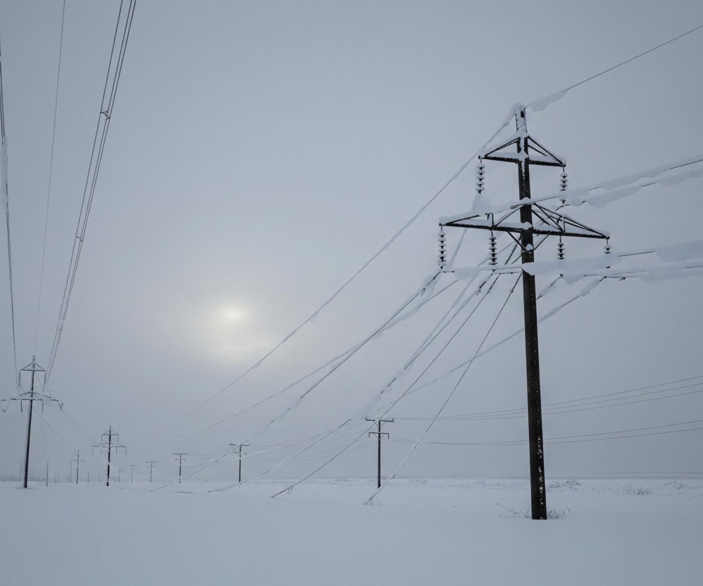 Zasypana śniegiem równina rozciąga się pod szarym, zachmurzonym niebem, na którym widać blednące słońce. Na pierwszym planie po prawej stronie wznosi się ciemnobrązowy słup wysokiego napięcia z ramionami pokrytymi grubą warstwą śniegu i lodu, co nadaje mu nieregularne kształty. Od słupa rozciągają się liczne, czarne linie przewodów, również obciążone śniegiem, tworząc falujące zwały lodu. W oddali, po lewej stronie i w centrum, widać szereg mniejszych słupów energetycznych, także pokrytych szronem, zanurzonych w śnieżnym krajobrazie, a za nimi rysuje się niewyraźna linia horyzontu i zalesiony fragment.