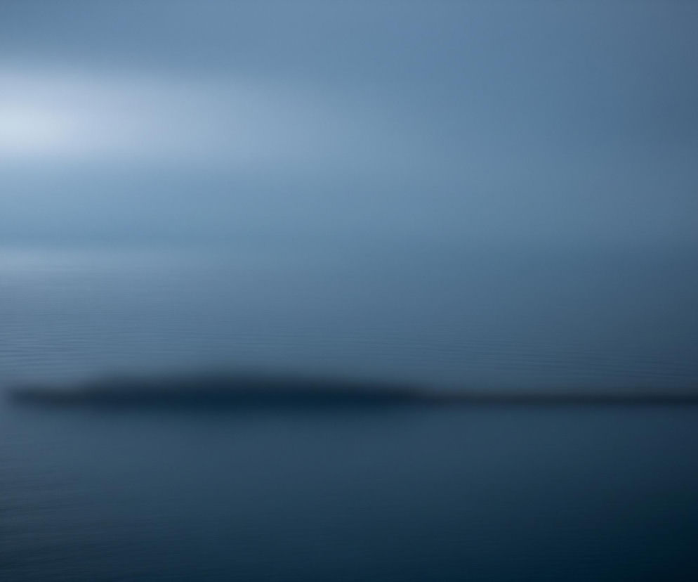 Rozległy, zamglony pejzaż wodny, zdominowany przez odcienie błękitu i szarości. W górnej lewej części kadru widoczne jest jasne rozbłysk światła, prawdopodobnie słońce, odbijające się od powierzchni wody. Horyzont jest niewyraźny, tworząc płynne przejście między niebem a wodą. Na wodzie, w środkowej części, dostrzec można zamazany, ciemniejszy kształt, przypominający wydłużoną, niską wyspę lub łódź, która płynie w kierunku prawej strony. Całość jest nieostra, co nadaje scenie eteryczny i tajemniczy charakter.