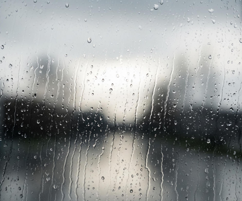 Na pierwszym planie widoczna jest mokra szyba pokryta licznymi kroplami deszczu, które spływają w dół, tworząc smugi. W tle, za szybą, zamazany miejski krajobraz w odcieniach szarości i bieli sugeruje zachmurzone niebo lub mglisty dzień. Po prawej stronie widoczny jest pionowy, ciemny element, który może być słupem lub framugą okna.