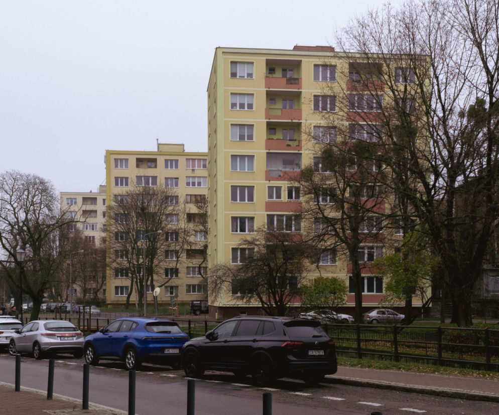 Ulica z rzędem zaparkowanych samochodów, a w tle kilka wysokich bloków mieszkalnych o jasnych, żółto-pomarańczowych elewacjach, symbolizujących wczesne budownictwo wysokich osiedli w Gdańsku. Więcej o ich historii przeczytasz na Architektura Murator Plus.