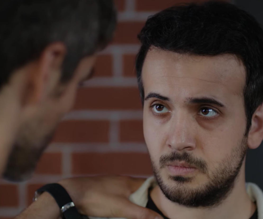 Poyraz w ujęciu z bliska, z zaniepokojonym wyrazem twarzy, rozmawiający z Mertem na tle ceglanej ściany. Zdjęcie ilustruje scenę, w której Poyraz wyznaje swoje uczucia do Nany, budząc zainteresowanie widzów serialu Dziedzictwo. Więcej informacji o serialu znajdziesz na Super Seriale.