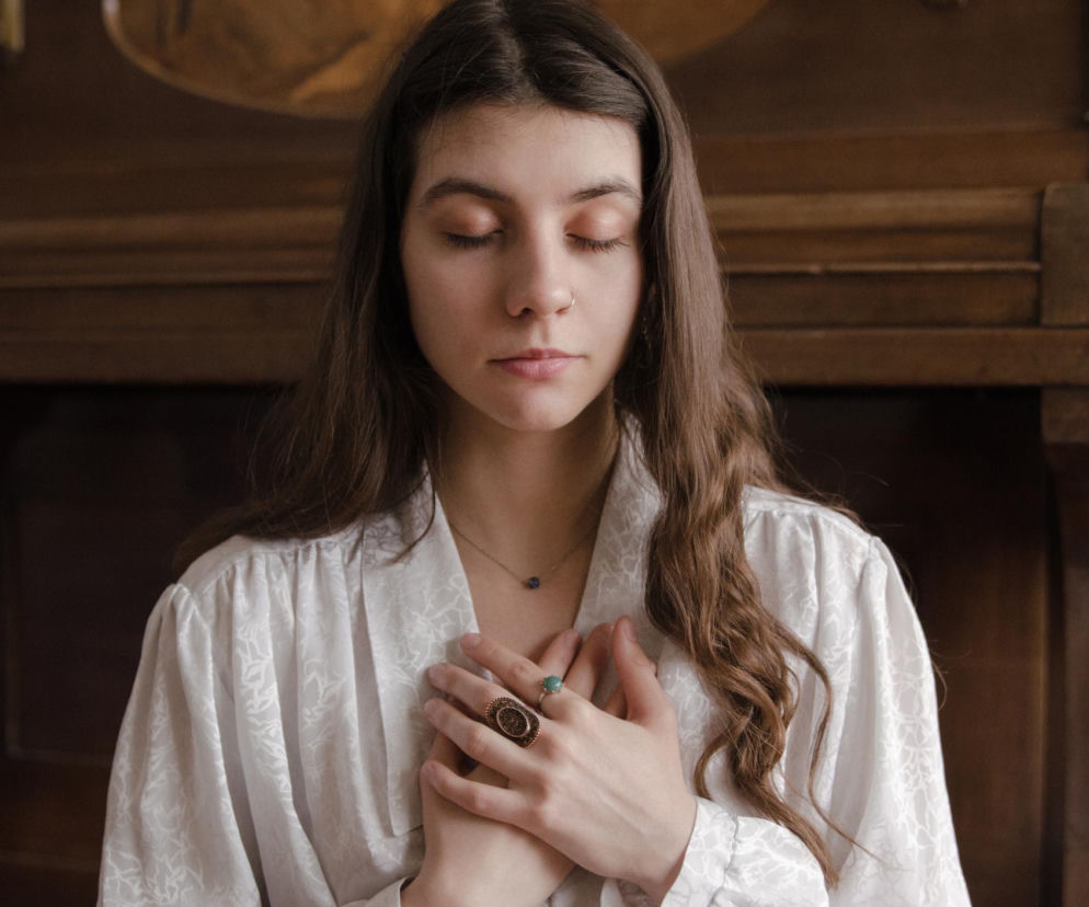 Młoda kobieta z długimi brązowymi włosami, w jasnej koszuli, z zamkniętymi oczami, trzymająca dłonie na klatce piersiowej w geście spokoju lub medytacji, co nawiązuje do technik wyciszania emocji i radzenia sobie z lękiem opisywanych na portalu Poradnik Zdrowie.