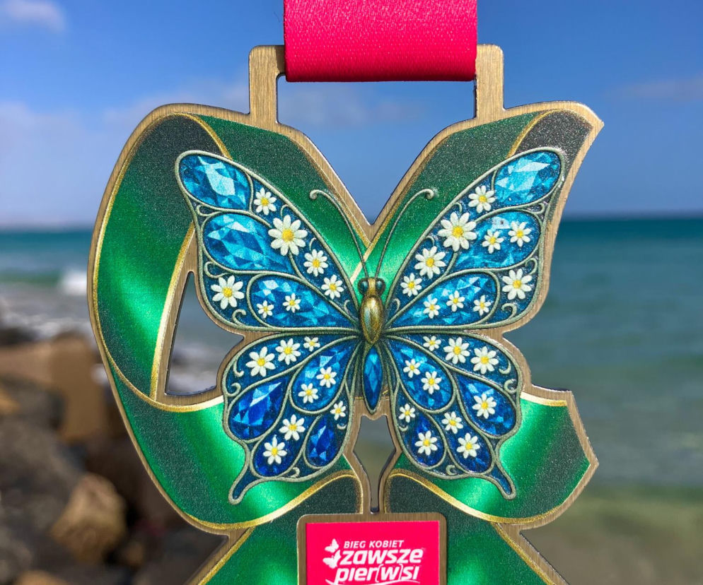 Medal Bieg Kobiet Zawsze Pier(w)si z motywem niebieskiego motyla i stokrotek na zielonym tle, zawieszony na różowej wstążce, z morzem i błękitnym niebem w tle. To symbol profilaktyki raka szyjki macicy, o której więcej przeczytasz na Poradnik Zdrowie.