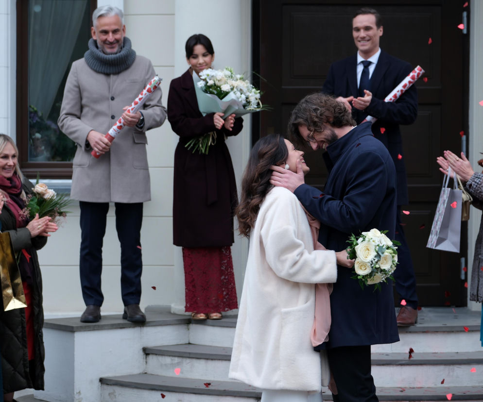 Para młoda Kasia (Paulina Lasota) i Mariusz (Mateusz Mosiewicz) całują się po ślubie, a Aneta (Ilona Janyst) i inni goście posypują ich płatkami w kształcie serc. Świadkowie i rodzina stoją na schodach budynku, gratulując młodym, których historia dostępna jest na Super Seriale.