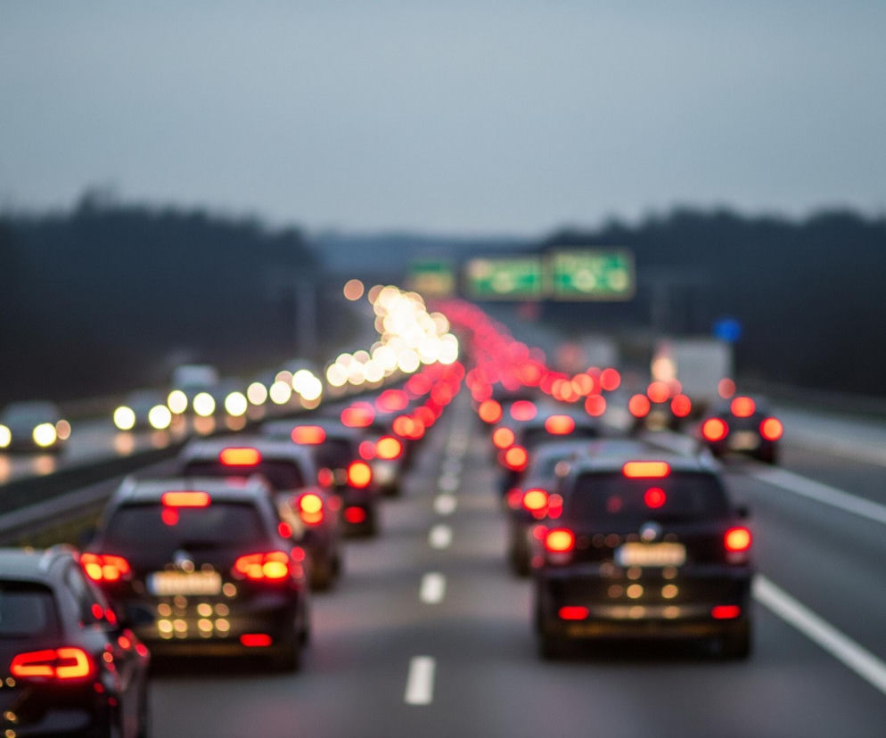 Pasy ruchu autostrady wypełnione są samochodami, ustawionymi w dwóch rzędach, z których lewy wydaje się być nieruchomy, a prawy ruszający. Tylne światła pojazdów tworzą ciąg czerwonych i pomarańczowych plam światła, a światła przednie na lewym pasie świecą na biało. Najbliżej widza znajduje się ciemny samochód osobowy, którego tablica rejestracyjna jest nieczytelna z powodu rozmycia. Horyzont dominuje szare, zachmurzone niebo, pod którym rozciąga się ciemny, rozmyty las.