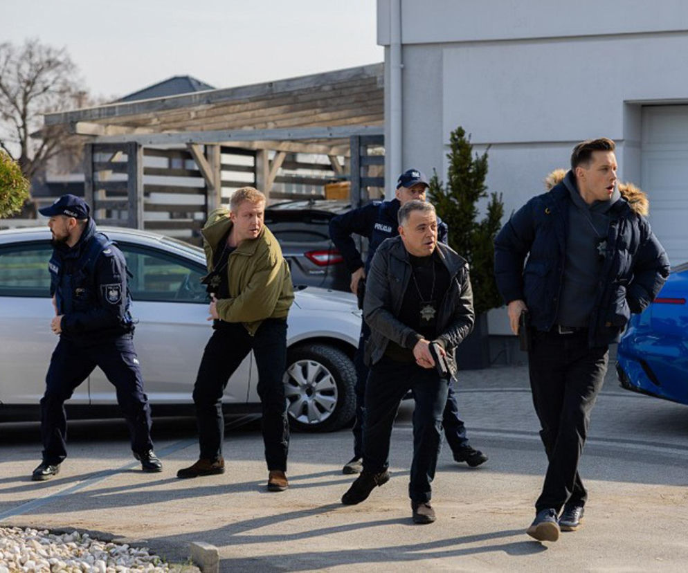 Grupa funkcjonariuszy policji w akcji, z bronią w rękach, w pozycji bojowej, otaczających budynek w tle. Scena dynamiczna, sugerująca napięcie i zagrożenie. Informacje o Pierwszej miłości znajdziesz na Super Seriale.