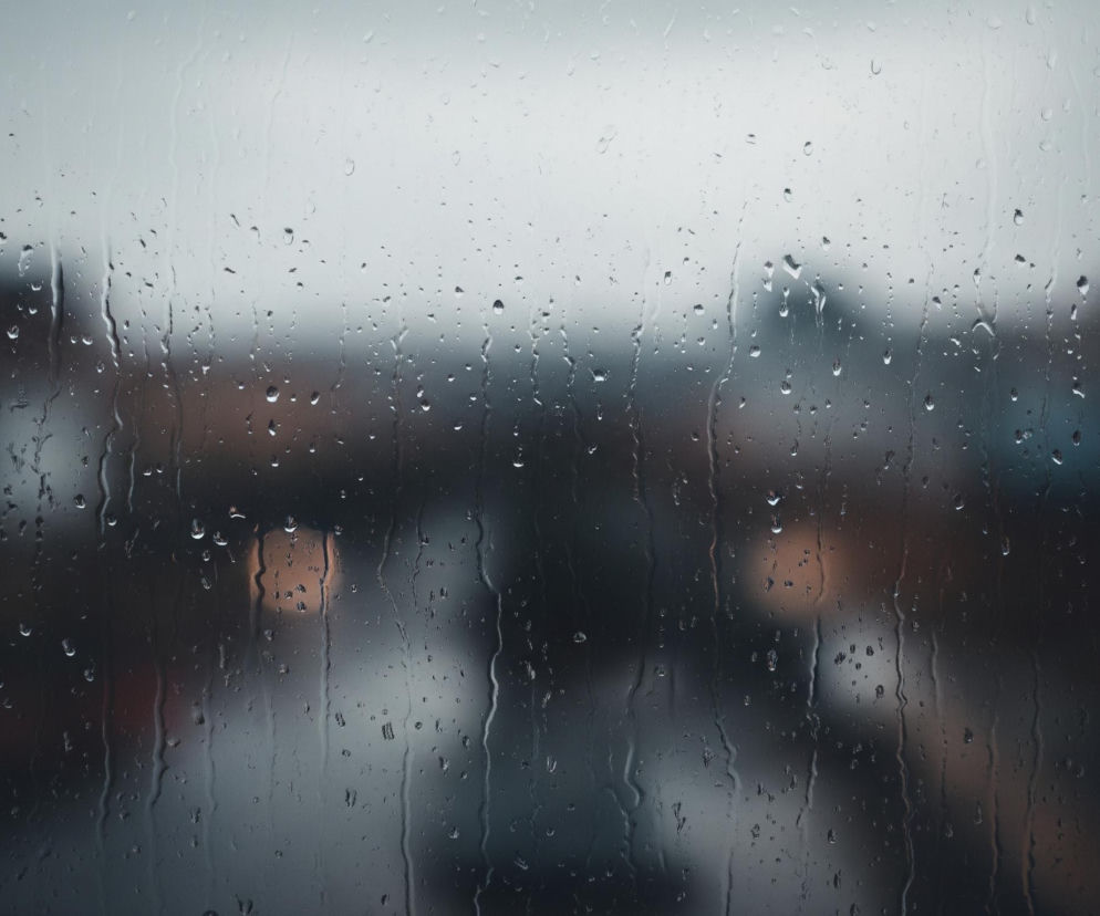 Obraz ukazuje szybę pokrytą licznymi kroplami deszczu, które spływają w dół, tworząc pionowe smugi. Tło za szybą jest rozmazane i ciemne, dominują odcienie szarości i granatu, z niewyraźnymi, jaśniejszymi plamami sugerującymi budynki lub światła w oddali. Górna część kadru jest jaśniejsza, przyjmując odcień bladego błękitu lub szarości, co wskazuje na pochmurne niebo.
