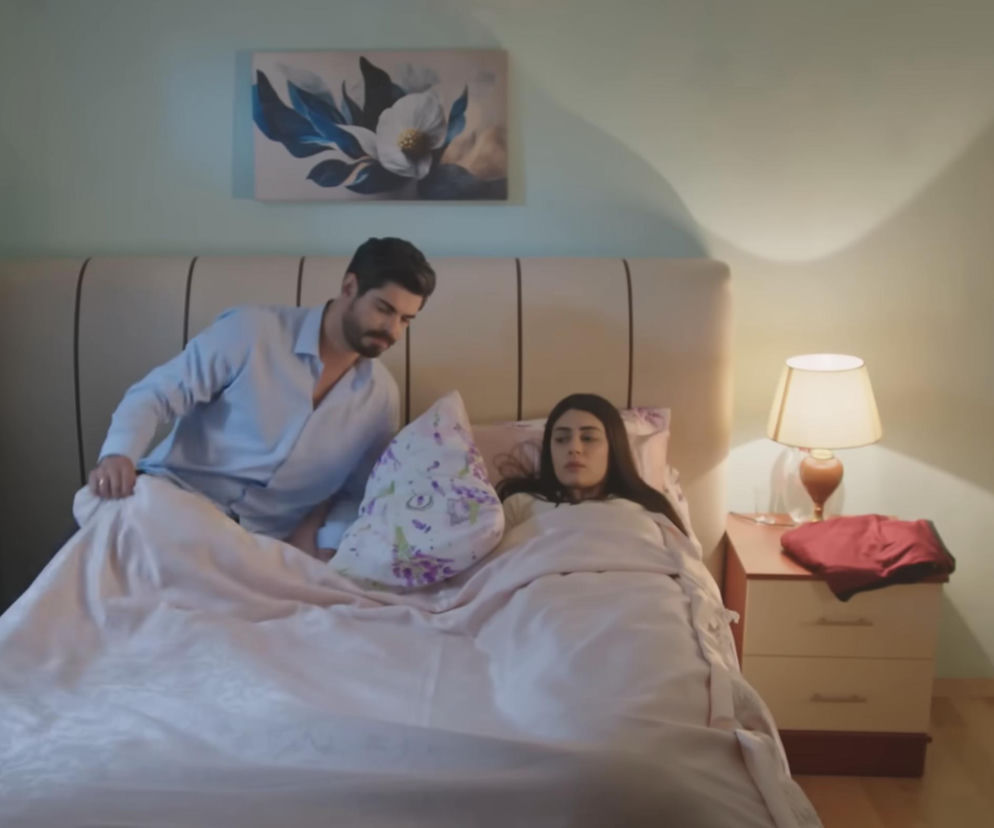 Cihan w jasnej koszuli kładący się do łóżka obok Hancer, która leży w nim w piżamie, w sypialni z jasnymi ścianami i obrazem kwiatów. Scena z tureckiego serialu Panna młoda o wspólnej nocy bohaterów, o której przeczytasz więcej na portalu Super Seriale.