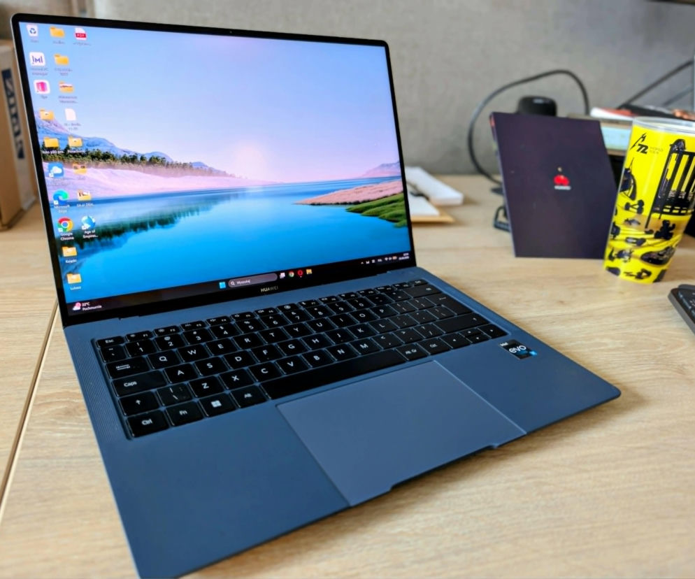 Laptop Huawei MateBook X Pro 2023 w kolorze ciemnoniebieskim, ukazany z otwartą klapą ekranu prezentującą jasny pulpit z tapetą przedstawiającą jezioro i góry. Widoczna klawiatura, duży touchpad oraz naklejka Intel Evo. W tle rozmyte biurowe otoczenie. Więcej o nim przeczytasz na Super Biznes.