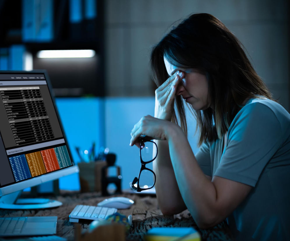 Kobieta cierpiąca na ból głowy i zmęczenie oczu, siedząca przy biurku z komputerem, przeciera skronie i trzyma okulary. To częsty sygnał ostrzegawczy cyfrowego zmęczenia wzroku, o czym przeczytasz więcej na Poradnik Zdrowie.
