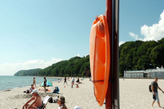 Pomarańczowa bojka ratunkowa wisi na słupku na plaży pełnej ludzi. O bezpieczeństwie nad wodą czytaj w Eska Olsztyn.
