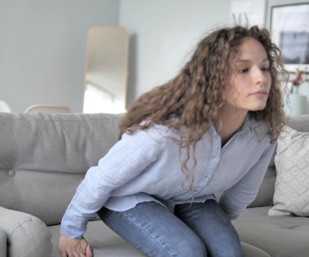 Kobieta o kręconych włosach, siedząca na szarej kanapie, próbuje wstać, podpierając się rękami o siedzisko. Ten gest może wskazywać na osłabienie mięśni nóg i niedobór witaminy D, o czym przeczytasz więcej na Poradnik Zdrowie.