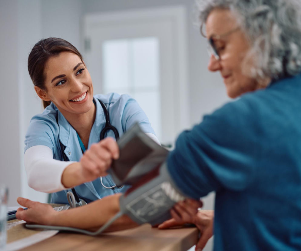 Uśmiechnięta młoda lekarka lub pielęgniarka w jasnoniebieskim uniformie medycznym mierzy ciśnienie krwi starszej kobiecie w okularach. Badanie domowe ciśnienia, o którym więcej przeczytasz na portalu Poradnik Zdrowie, to klucz do monitorowania nadciśnienia.
