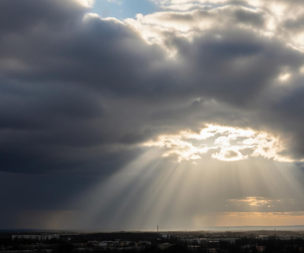Słońce przebija się przez gęste, ciemnoszare chmury, tworząc jasne smugi światła skierowane ukośnie w dół. Centrum nieba jest rozświetlone, kontrastując z otaczającymi, burzowymi obłokami, a fragmenty błękitnego nieba są widoczne w górnej części kadru. Poniżej chmur rozciąga się miejski krajobraz z licznymi budynkami i drzewami, zlewający się w ciemną, rozmytą sylwetkę, nad którą widać delikatną mgiełkę. Horyzont na prawo jest jaśniejszy, sugerując oddalenie się burzowych warunków.