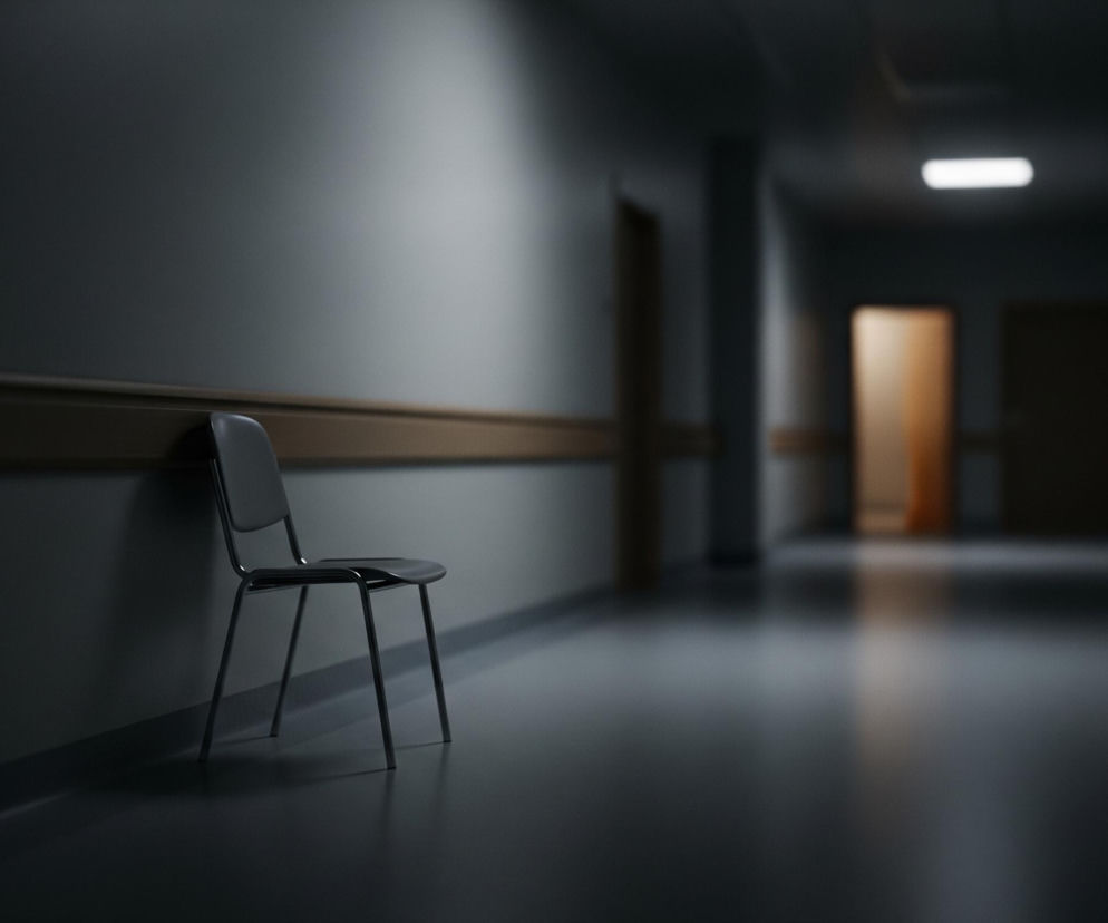 Wąski korytarz, prawdopodobnie w budynku publicznym, jest pogrążony w półmroku. Po lewej stronie, blisko ściany, stoi pojedyncze, czarne krzesło o prostej konstrukcji. Na jasnoszarych ścianach, na wysokości około bioder, biegnie pozioma, drewniana lub drewnopodobna listwa, a na suficie korytarza widoczne jest jedno prostokątne, jarzeniowe światło. Dalej w głąb korytarza, po obu stronach, widoczne są drzwi, z których jedne, po prawej stronie, są częściowo otwarte i wydobywa się z nich ciepłe, żółtopomarańczowe światło, kontrastujące z chłodną tonacją reszty przestrzeni.