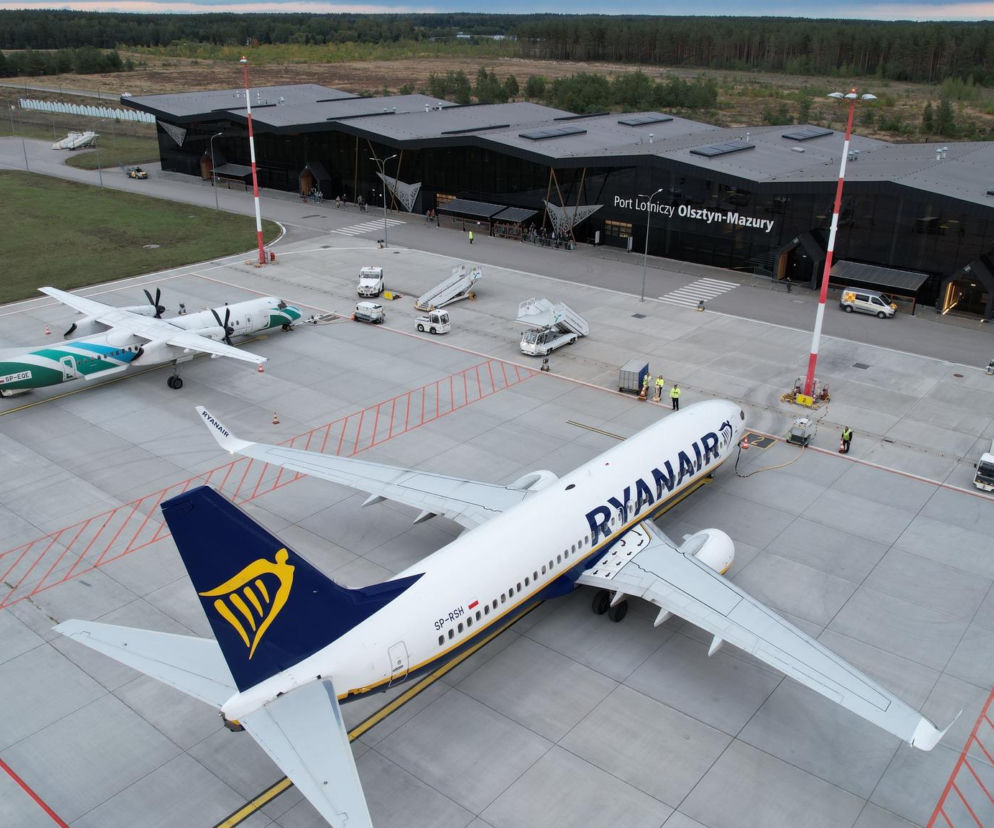 Widok z lotu ptaka na Port Lotniczy Olsztyn-Mazury z białym samolotem Ryanair i mniejszym turbośmigłowym na płycie postojowej. Terminal z czarnym dachem i panelami fotowoltaicznymi w tle otacza las. Więcej o inwestycjach lotniska na Architektura Murator Plus.